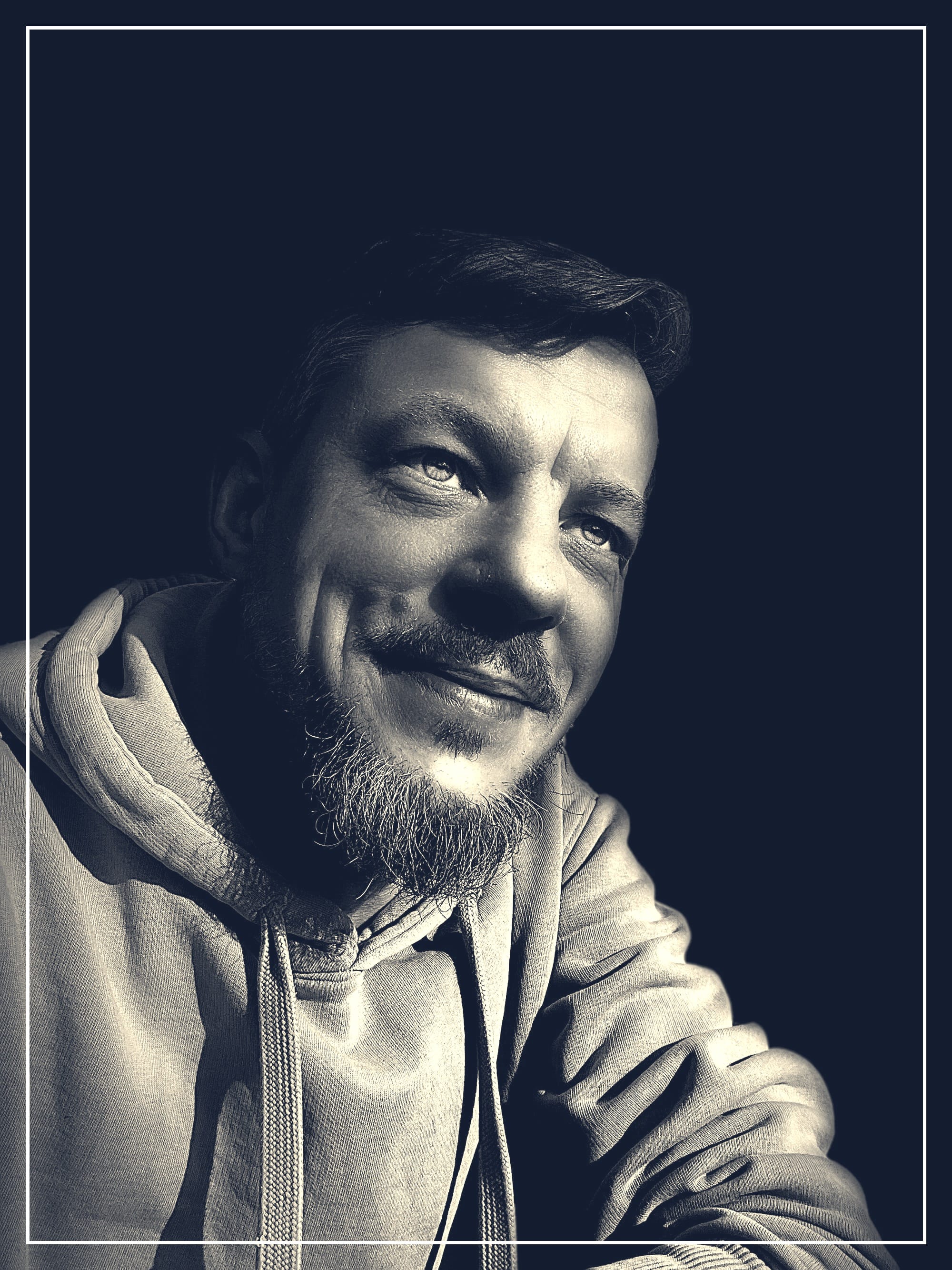 Ein Mann mit kurzem Haar und Bart sitzt in weichem Licht vor schwarzem Hintergrund. Er trägt einen Hoodie, lächelt leicht und blickt nach oben rechts, als würde er nachdenken oder einen stillen Moment genießen.